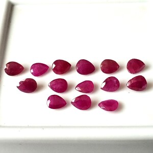 May include: A collection of 14 pear-shaped ruby gemstones, each with a deep red hue and a faceted cut. The gemstones are arranged on a white surface.
