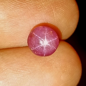 May include: A round, cabochon-cut star ruby gemstone featuring a deep magenta-red hue. The stone displays a distinct six-rayed asterism effect, where a bright white star pattern radiates from the center of the polished, smooth, and domed surface.