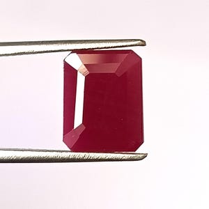 May include: A deep red emerald cut gemstone, held by a pair of tweezers.