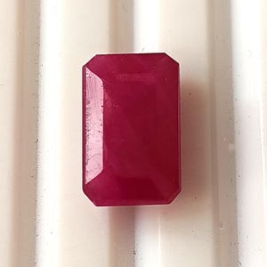 May include: A rectangular, faceted ruby gemstone with a deep red hue. The gemstone has an emerald cut with beveled edges and a polished surface. The ruby is set against a white background.