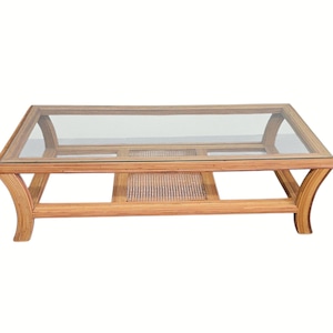 May include: A rectangular coffee table with a clear glass top and a natural rattan frame. The table features curved legs and a woven rattan shelf. The overall design is light brown and airy, suitable for a living room.