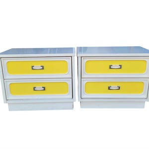 May include: A pair of white bedside tables, each featuring two drawers with bright yellow front panels. The drawers are equipped with rectangular white handles set against dark metallic backplates, providing a clear contrast.