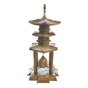 May include: A brass pagoda-style sculpture featuring a multi-tiered roof structure, four vertical pillars, and a central hanging bell. The base is decorated with embossed dragon motifs and a central symbol, all finished in a metallic gold tone.