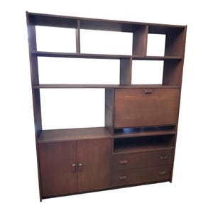 May include: A tall, dark brown wooden shelving unit featuring multiple open storage compartments, a drop-down cabinet door, two lower cabinet doors with small handles, and two stacked drawers with matching hardware on the right side.