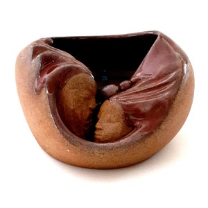 Puede incluir: Un cuenco de cerámica hecho a mano con un diseño único. El cuenco presenta dos caras esculpidas, una a cada lado, con un interior marrón oscuro. El exterior es de color marrón cálido con acentos de un esmalte marrón rojizo más oscuro.