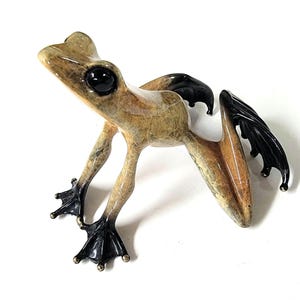 May include: A bronze frog sculpture with black eyes and webbed feet. The frog has black wings and a textured, mottled brown and tan body. The sculpture is posed as if leaping forward, with its legs extended.