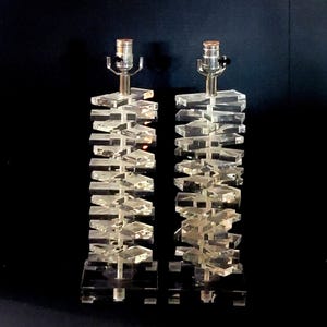 May include: Two clear acrylic table lamps with a stacked geometric design. Each lamp has a silver-toned base and a light fixture at the top. The lamps are set against a black background.