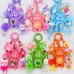 Op de afbeelding: Zes Care Bears sleutelhangers met plastic berenfiguren met schudbuiken, kleurrijke kralen, bedels en pompons. De beren zijn paars, roze, blauw, rood, groen en oranje, elk met unieke symbolen en diverse bijgevoegde decoratieve elementen.