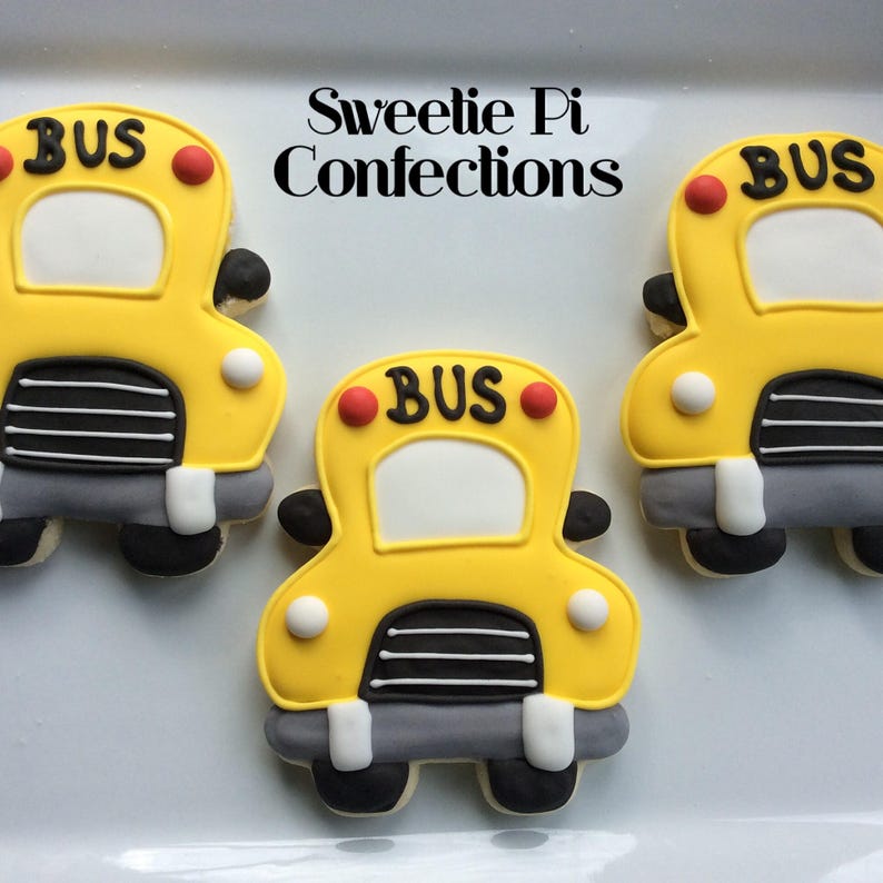 May include: Three yellow school bus sugar cookies decorated with black and white details. The cookies have red circles for headlights and the word "BUS" written in black on the front.
