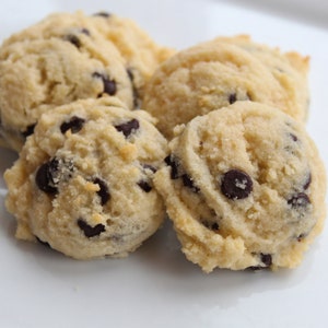 May include: Four soft, light brown cookies with chocolate chips on a white surface.