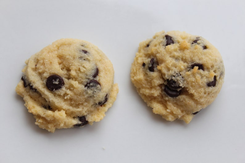 May include: Two light brown chocolate chip cookies on a white surface. The cookies are slightly misshapen and have a few chocolate chips visible.
