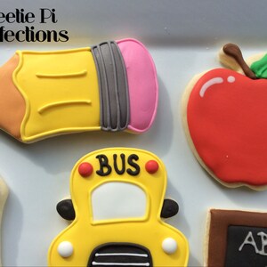 May include: A set of five decorated sugar cookies with a back to school theme. The cookies are shaped like a pencil, an apple, a school bus, a crayon, and a chalkboard. The cookies are decorated with colorful icing and have details like "BUS" on the school bus and "ABC" on the chalkboard.