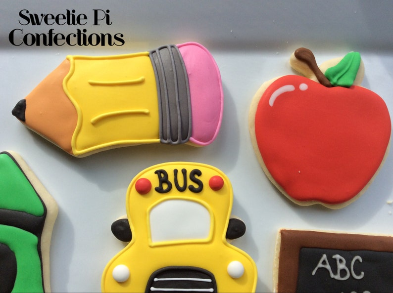 May include: A set of five decorated sugar cookies with a back to school theme. The cookies are shaped like a pencil, an apple, a school bus, a crayon, and a chalkboard. The cookies are decorated with colorful icing and have details like "BUS" on the school bus and "ABC" on the chalkboard.