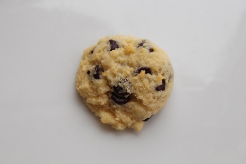 May include: A single, round, light brown cookie with chocolate chips. The cookie is slightly flattened and has a rough texture.