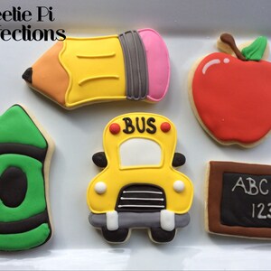 May include: Five decorated sugar cookies with a back to school theme. The cookies are shaped like a pencil, apple, crayon, school bus, and a chalkboard. The chalkboard has the letters ABC and the numbers 123 written on it.
