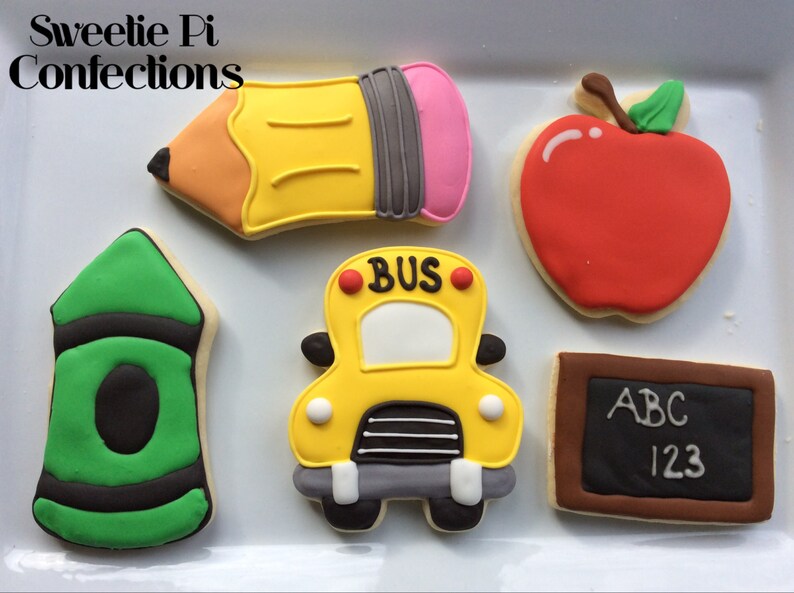 May include: Five decorated sugar cookies with a back to school theme. The cookies are shaped like a pencil, apple, crayon, school bus, and a chalkboard. The chalkboard has the letters ABC and the numbers 123 written on it.