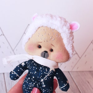 May include: A small plush toy in the shape of a sheep wearing a white fuzzy hat with pink ears and a blue and white starry patterned outfit. The toy is being held in a hand.