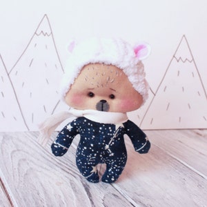 May include: A plush toy bear wearing a white fluffy hat with pink ears and a white scarf. The bear is wearing a blue and white outfit with a star pattern. The bear is standing on a white wooden surface.