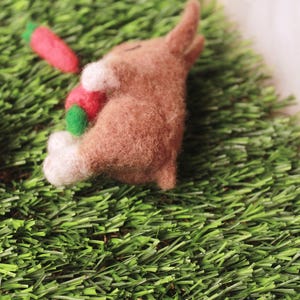 May include: A brown felt bunny rabbit laying on green grass with a red and green carrot in its paws.