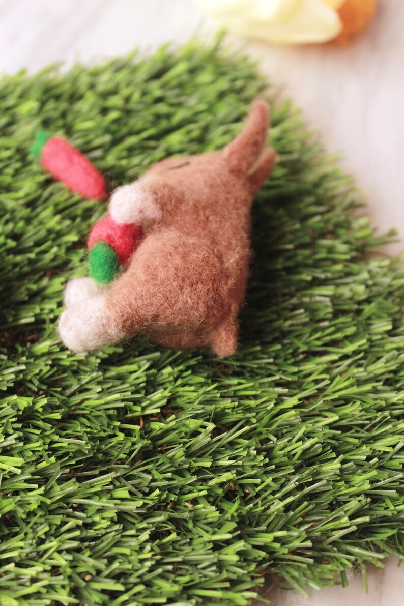 May include: A brown felt bunny rabbit laying on green grass with a red and green carrot in its paws.