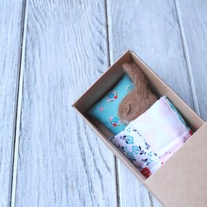May include: A small brown felt animal is sleeping in a cardboard box. The animal is lying on a blue and white floral pillow with a pink and white floral blanket.