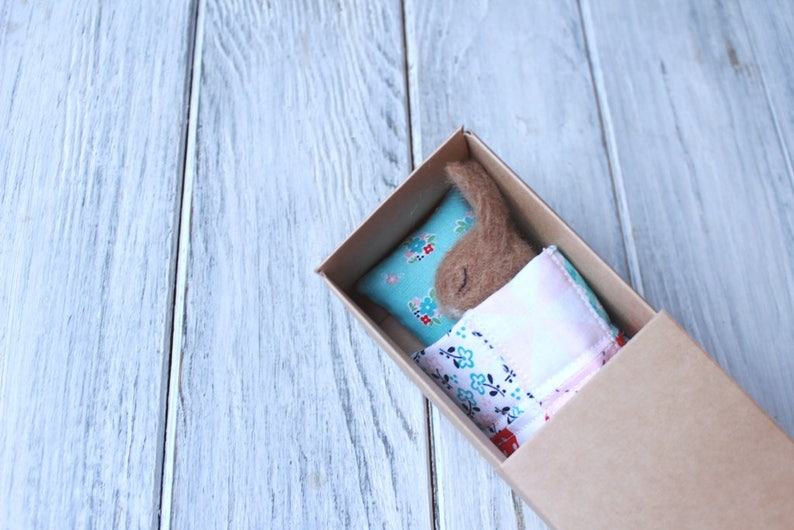 May include: A small brown felt animal is sleeping in a cardboard box. The animal is lying on a blue and white floral pillow with a pink and white floral blanket.