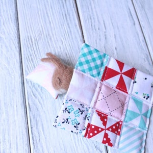 May include: A brown and pink felt bunny toy is tucked into a patchwork quilt made of various fabrics with different patterns, including gingham, polka dots, and floral prints.