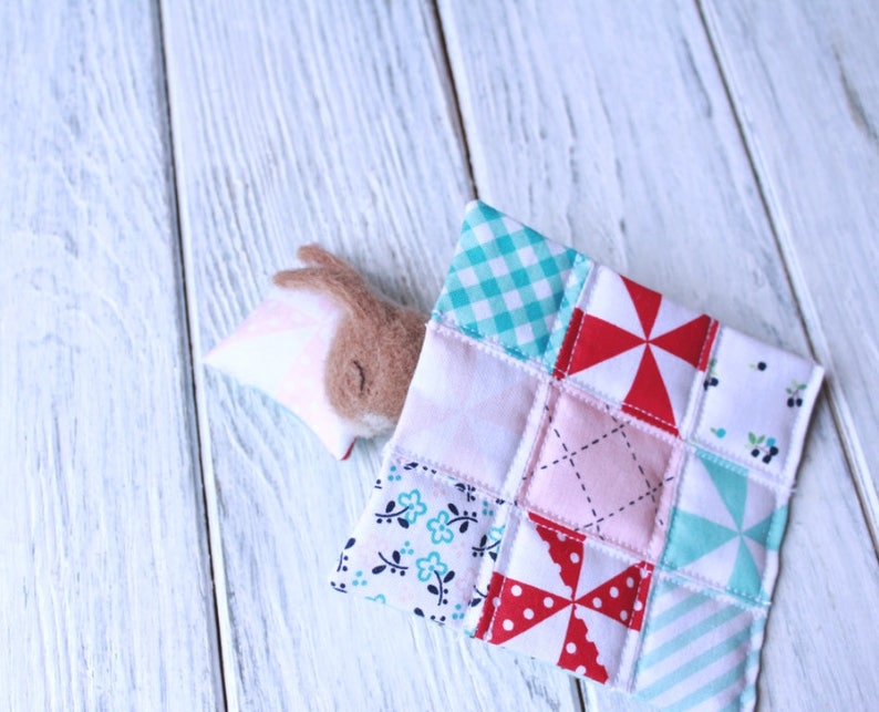 May include: A brown and pink felt bunny toy is tucked into a patchwork quilt made of various fabrics with different patterns, including gingham, polka dots, and floral prints.