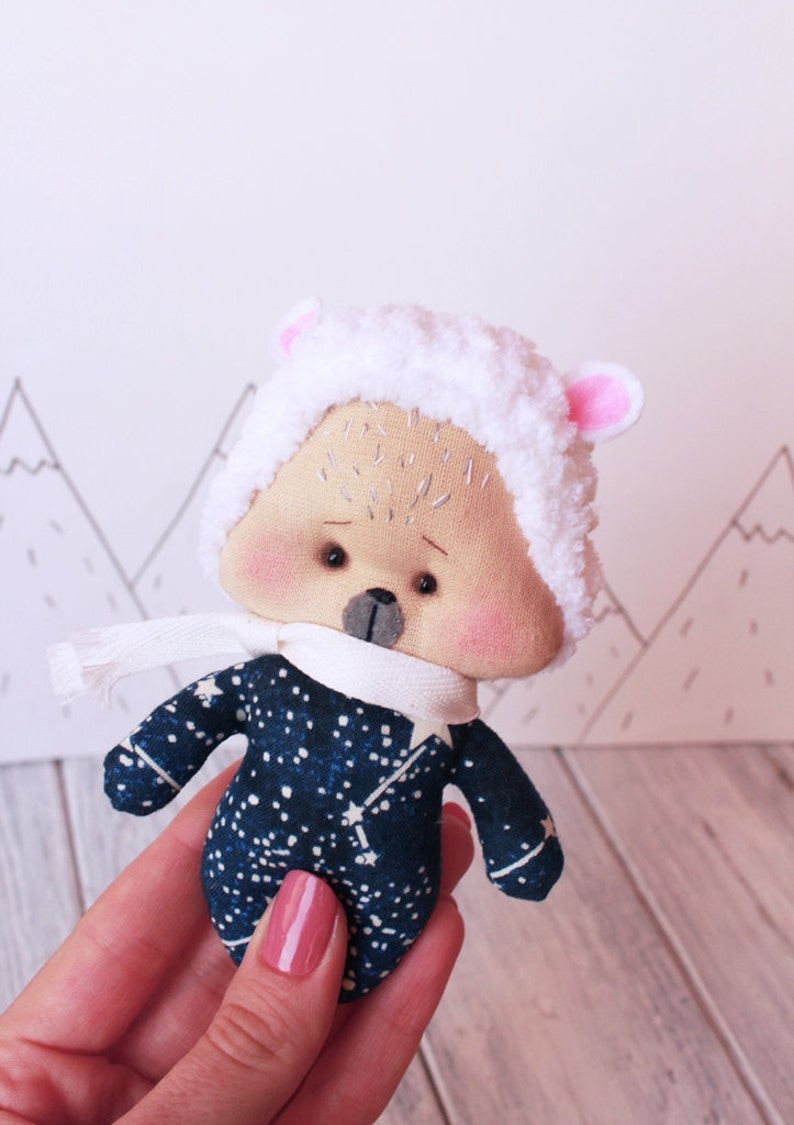 May include: A small plush toy in the shape of a sheep wearing a white fuzzy hat with pink ears and a blue and white starry patterned outfit. The toy is being held in a hand.