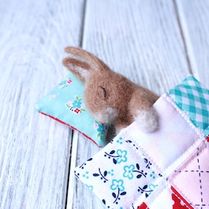 May include: A brown felt bunny is sleeping in a patchwork quilt with a blue and white floral pillow. The quilt is made of different fabrics in various colors and patterns.