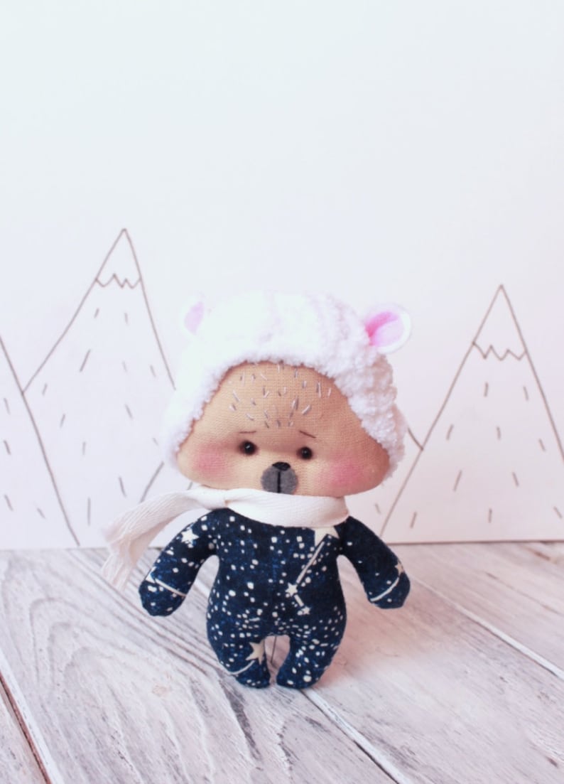 May include: A plush toy bear wearing a white fluffy hat with pink ears and a white scarf. The bear is wearing a blue and white outfit with a star pattern. The bear is standing on a white wooden surface.