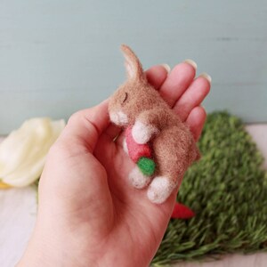 May include: A small brown felt bunny with white feet is holding a red and green felt carrot in its paws. The bunny is lying on its side with its eyes closed, as if it is sleeping.