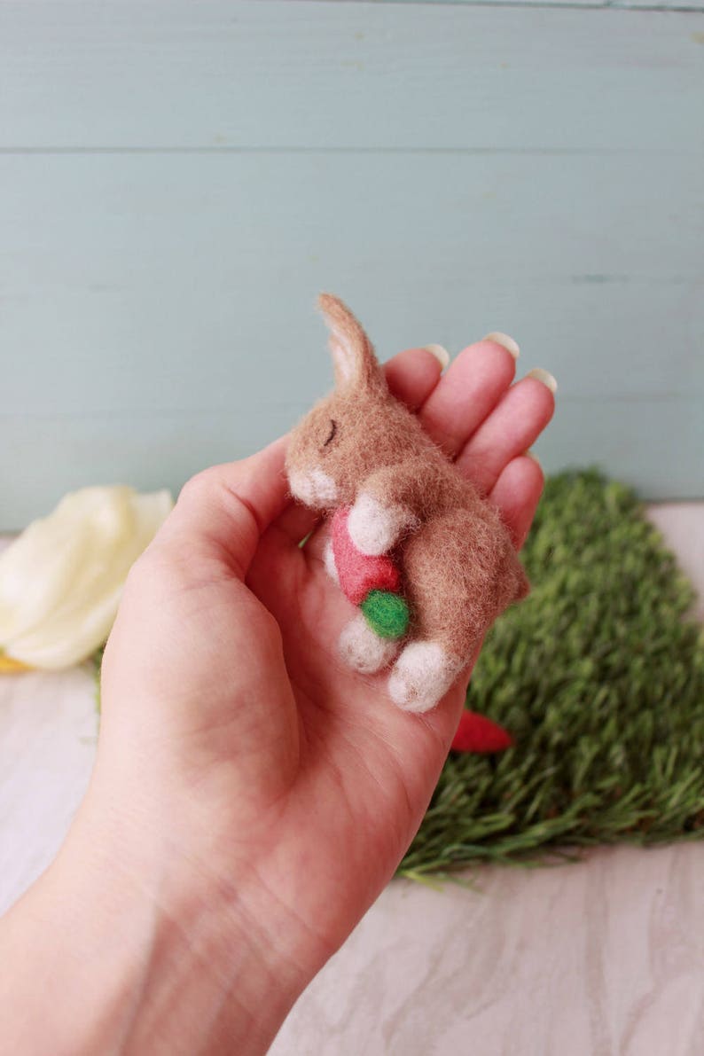 May include: A small brown felt bunny with white feet is holding a red and green felt carrot in its paws. The bunny is lying on its side with its eyes closed, as if it is sleeping.