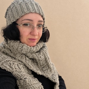 May include: A light gray crocheted beanie hat and a matching long crocheted scarf featuring a textured cable knit pattern. The set is crafted from a solid light gray yarn, showcasing intricate loops and stitches throughout the fabric.