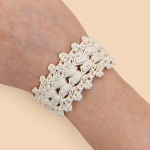May include: A cream-colored crochet bracelet featuring a repeating pattern of thick, vertical cord-like stitches bordered by delicate, scalloped edges. The design is crafted from a continuous thread, creating a textured, woven appearance.