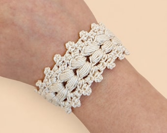 May include: A cream-colored crochet bracelet featuring a repeating pattern of thick, vertical cord-like stitches bordered by delicate, scalloped edges. The design is crafted from a continuous thread, creating a textured, woven appearance.