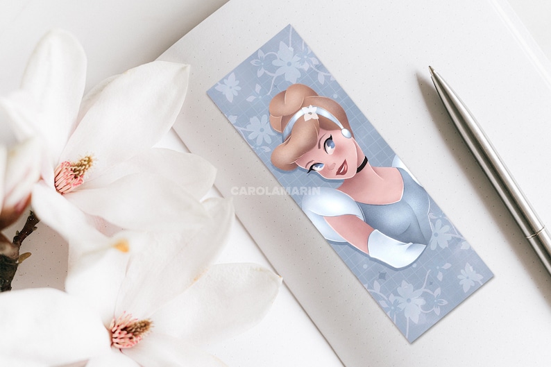 Op de afbeelding: Een blauwe en witte bladwijzer met een portret van Assepoester, een Disney-prinses, in een lichtblauwe jurk en een wit hoofdband. De achtergrond is lichtblauw met een wit bloemenpatroon.