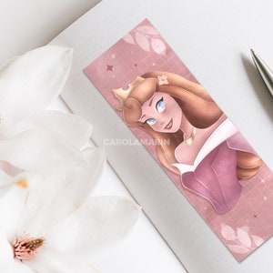 Op de afbeelding: Een roze en gouden bladwijzer met een cartoonillustratie van Doornroosje, een Disney-prinses, met een kroon en een roze jurk. De achtergrond is een roze en wit rasterpatroon met witte bladeren.