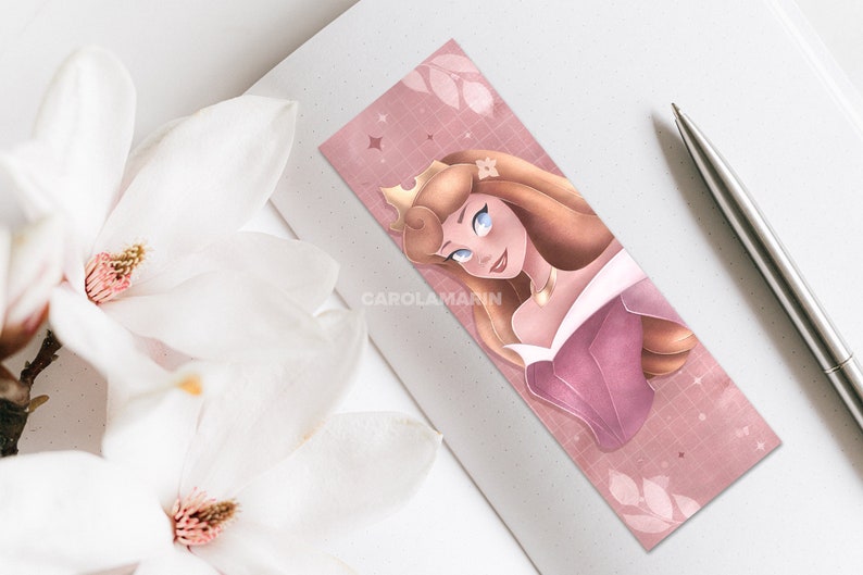 Op de afbeelding: Een roze en gouden bladwijzer met een cartoonillustratie van Doornroosje, een Disney-prinses, met een kroon en een roze jurk. De achtergrond is een roze en wit rasterpatroon met witte bladeren.
