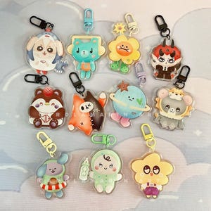 May include: Collection of colorful acrylic keychains featuring various cartoon characters. Designs include a bear with a basketball, a cat, a devil, a bear with headphones, a flower, a planet, a mouse, a dog, a baby, and a cloud.