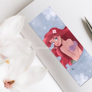 Op de afbeelding: Een bladwijzer met een cartoonillustratie van Ariel, de Kleine Zeemeermin, met rood haar, een paarse schelpbeha en een groene staart. De achtergrond is lichtblauw met witte bloemen en sterren.