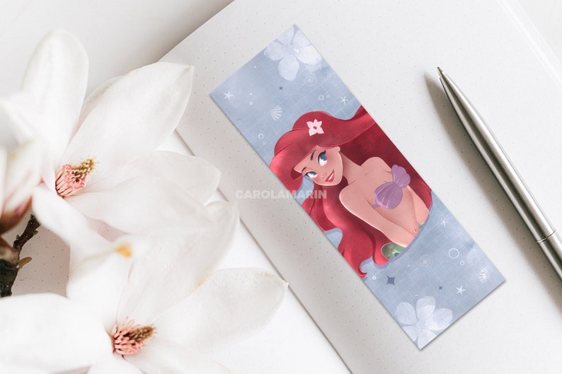 Op de afbeelding: Een bladwijzer met een cartoonillustratie van Ariel, de Kleine Zeemeermin, met rood haar, een paarse schelpbeha en een groene staart. De achtergrond is lichtblauw met witte bloemen en sterren.