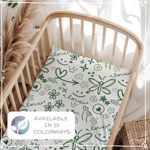 Könnte beinhalten: Ein weißes Spannbettlaken für das Kinderbett mit einem grünen Kritzelmuster aus Schmetterlingen, Herzen, Sternen und Blumen. Das Laken befindet sich in einem hellen Holzbett. Der Text "AVAILABLE IN 19 COLORWAYS" steht in einer weißen Box.