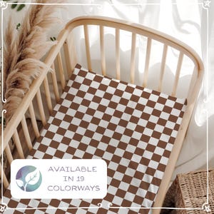 Può includere: Un lenzuolo a quadretti marrone e bianco per culla all'interno di una culla di legno. Il lenzuolo presenta un motivo ripetuto di quadrati marroni su sfondo bianco. L'immagine include il testo "AVAILABLE IN 19 COLORWAYS".