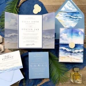 May include: Wedding invitation suite with a beach theme. The invitation features a watercolor painting of the ocean and sky. The suite includes an invitation, RSVP card, details card, and envelope. The color scheme is blue and white, with gold accents.