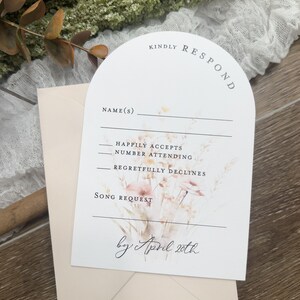 May include: A white RSVP card with a floral design and the text "KINDLY RESPOND" in a curved arch. The card has fields for the guest's name, RSVP options, and a song request. The text "by April 20th" is written in cursive at the bottom of the card.