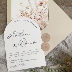 May include: A white wedding invitation with a floral design and a wax seal. The invitation reads "Please join us to celebrate Andrea & Robnah" and includes the date, time, and location of the wedding. The text "Reception to Follow" is also included.