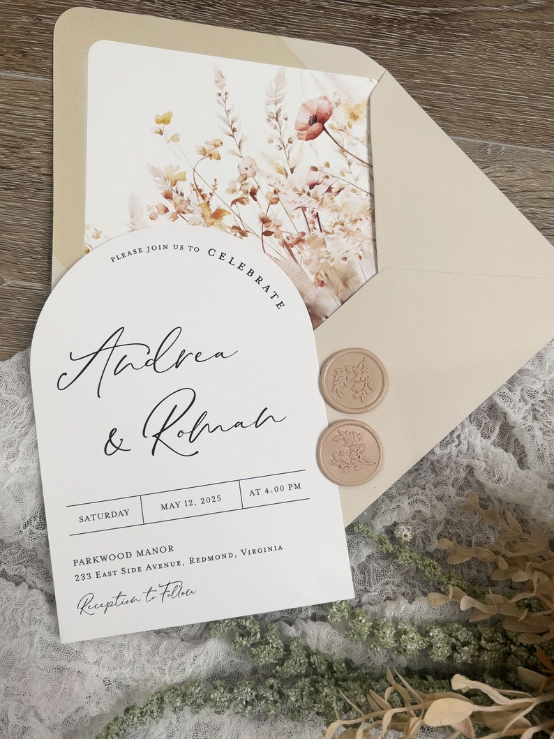 May include: A white wedding invitation with a floral design and a wax seal. The invitation reads "Please join us to celebrate Andrea & Robnah" and includes the date, time, and location of the wedding. The text "Reception to Follow" is also included.
