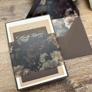 May include: A wedding invitation suite featuring a dark brown envelope with a floral patterned liner, a cream-colored card, a tan card, and a dark brown RSVP card with the text Kindly Respond, all held together by a translucent floral belly band.