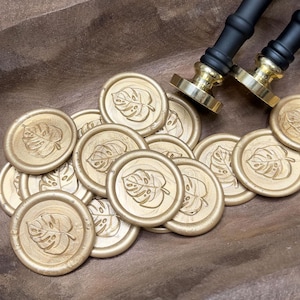 May include: Gold wax seals with a monstera leaf design are scattered in a wooden bowl. Two black and gold wax seal stamps are also in the image. The seals are round and have a raised leaf pattern. The bowl is a warm brown color.